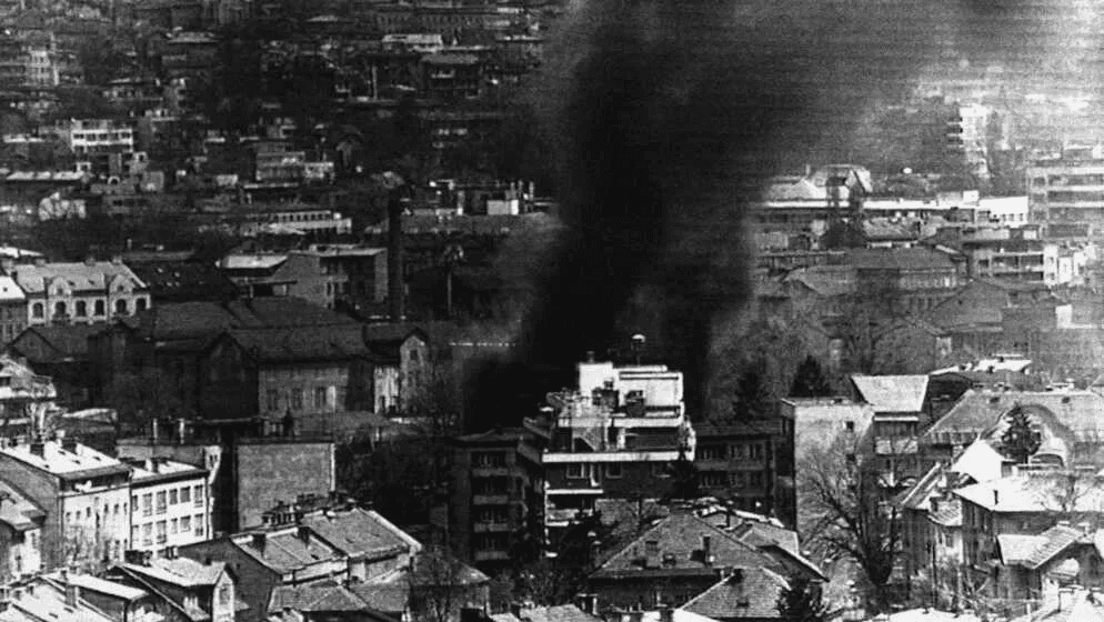 Black-and-white photograph of smoke rising from a shelled building in downtown Sarajevo in 1992.
